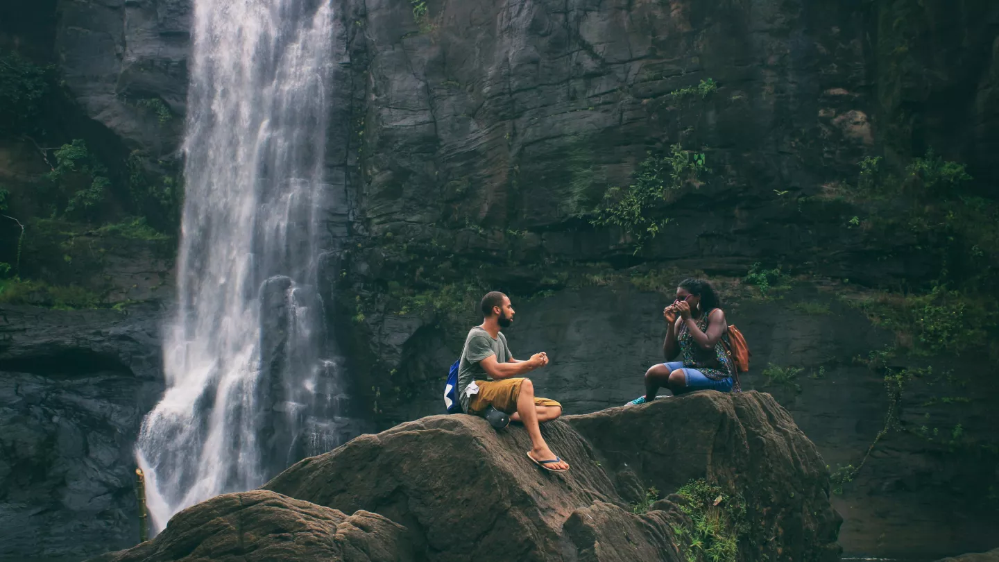 Two people sit on rocks beside a cascading waterfall